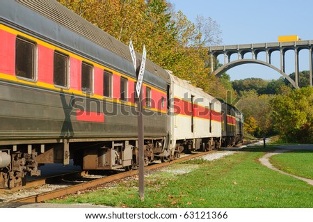 A scenic passenger train nears a high arch bridge in a rural area