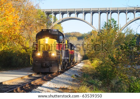 An oncoming passenger train under a high arch bridge in a scenic area