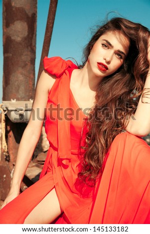 Fashion portrait of classy brunette in red shiny dress and long wavy hair . vertical shot , outdoors. luxury photography.