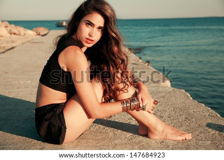 Beautiful fashionable model with long hair sitting on the beach in black shorts and t shirt . Horizontal evening shot.