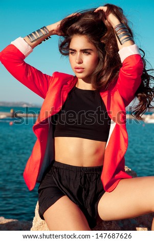 Fashionable brunette woman with long hair in summer outfits and red Jacket  posing on a sunny day by the sea . outdoors shot, soft sunny colors . vertical shot.