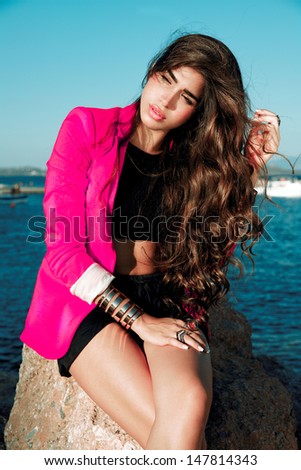 Fashionable brunette woman with long hair in summer outfits and pink Jacket  posing on a sunny day near a sea