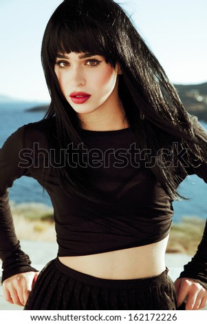 Vogue style brunette in Asian style by the sea. Fashion art. Woman with long black hair and black outfits. Blue sky backgrounds. Bright make up face .Red lips . sexy look. Street style. Vertical shot.