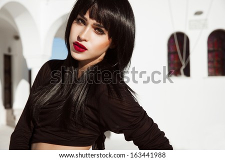 Attractive young brunette in black outfits and shine hair. Bright make up with red lips. Shine straight hair. Sexy female in front of a white building. Santorini day Outdoors shot. horizontal.