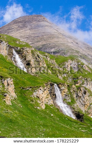 green slopes with two waterfalls and a mountain peak with cloud veils around and a blue sky above