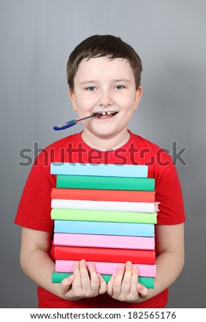 Boy with a pen in his mouth holding a stack of books, gray background.
