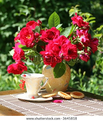 In a garden there is a little table with a bouquet of roses and cup of tea. On plates lay cookies and a red currant.