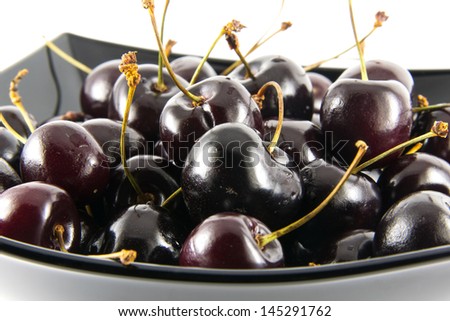 Fresh ripe black cherries with sticks in a black bowl. Closeup on a white background