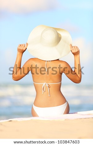 Travel vacation woman on summer holiday on beach holding her summer hat sitting in the sand enjoying ocean. Back view of beautiful girl in white bikini.