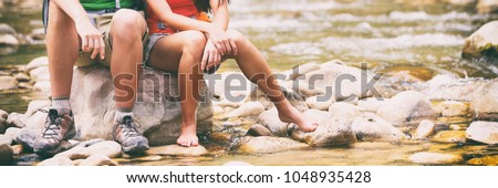 Hiking couple hikers resting on rock putting barefoot feet in water. Woman and man hiker sitting by river creek, boots footwear legs closeup. Young couple trekking, relaxing after hike, USA travel.