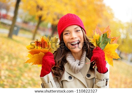 Fall woman happy and bliss in autumn city forest park holding colorful fall leaves smiling happy and joyful wearing tuque and knit gloves. Pretty multiracial Asian and Caucasian girl model.