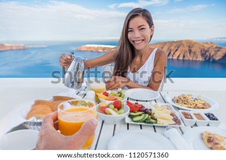 Couple eating breakfast at luxury resort - honeymoon Europe travel vacation, Santorini, Greece. Asian woman drinking coffee on outdoor hotel restaurant, Mediterranean sea view. Healthy food brunch.