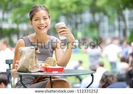 Young business woman on lunch break in City Park drinking coffee and eating sandwich. Happy smiling multiracial young businesswoman in Bryant Park, New York City, USA.