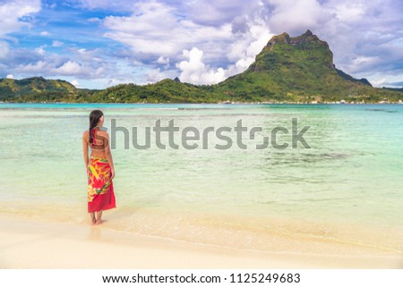Tahiti luxury travel beach vacation woman walking in polynesian cover-up skirt beachwear on idyllic paradise island in French Polynesia. Red traditional outfit,, bikini and flower girl.
