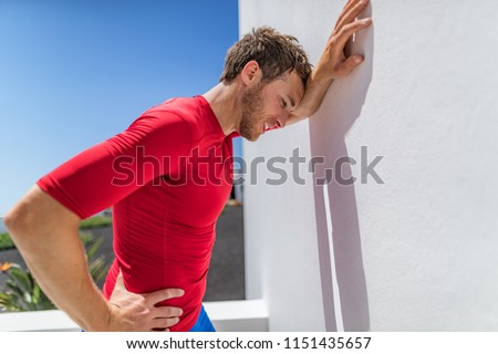 Tired athlete runner man exhausted leaning on wall of fatigue breathing hard after difficult exercise. Fitness person sweating of sun stroke, migraine, heat exhaustion muscle back pain or cramps.