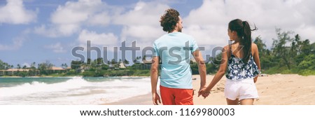 Happy young couple holding hands walking on Hawaii beach during holidays travel. People tourists in their 20s casual enjoying summer living. Banner panorama portrait.