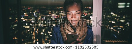 Woman texting instant messaging text on mobile phone online at night - bright light from screen illuminating young Asian girl in London city background banner panorama.