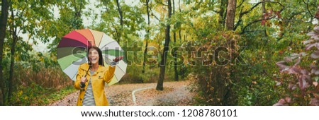 Autumn woman walking in rain under umbrella. Happy asian girl enjoying park forest outdoors looking up joyful on rainy fall day wearing yellow raincoat outside in nature. Multiracial Asian girl.