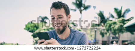 Smartphone man using mobile phone texting sms text message outside in summer background. Young people lifestyle banner panorama. Happy casual guy smiling holding cellphone using app for social media.