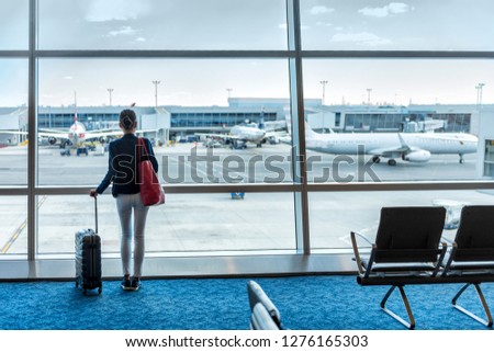 Traveler businesswoman waiting for delayed flight at airport lounge standing with luggage watching tarmac at airport window. Woman at boarding gate before departure. Travel lifestyle.