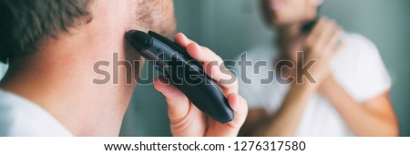 Man shaving beard using electric trimmer shaver. Male beauty grooming banner panorama. Home lifestyle young person looking at bathroom mirror trimming hair on neck.