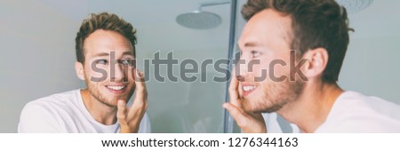 Man putting cream on face in bathroom mirror banner panorama. Guy applying anti-aging skin care product moisturizer under eyes for facial treatment. Male beauty morning routine at home lifestyle.