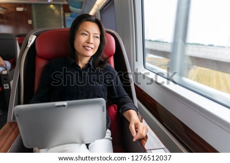 Business trip Asian woman working on laptop on train commute travel lifestyle. Middle aged chinese businesswoman smiling looking out window in contemplation.