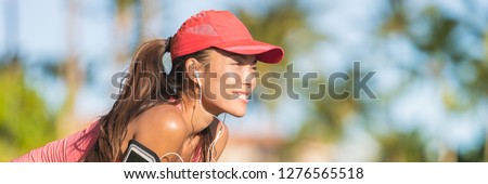 Summer running healthy active woman runner listening to music on phone sport armband with headphones earphones on city street. Happy fitness Asian jogging girl with sports cap ready banner panorama.