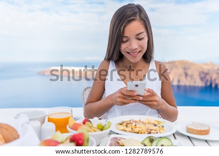 Food selfie social media vlogging girl taking photo with phone of breakfast plate using cellphone app. Luxury travel vacation lifestyle. Beautiful model in resort in Santorini, Greece, Europe.