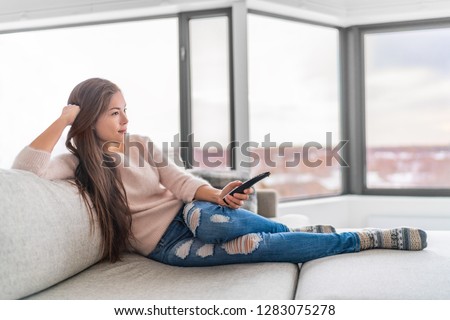Woman watching TV at home sitting on sofa in modern apartment. Young casual Asian girl enjoying TV show holding remote. Comfort living lifestyle.