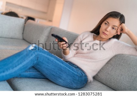Bored woman watching tv alone in apartment sitting on sofa holding remote pensive. Asian girl tired at home relaxing on couch having nothing to do but watching television.