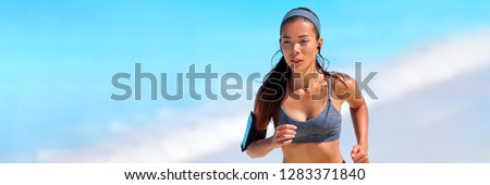 Healthy fit active Asian woman on beach run wearing technology wearable tech device smartphone holder and earphones listening to music motivation on summer workout -banner panorama.