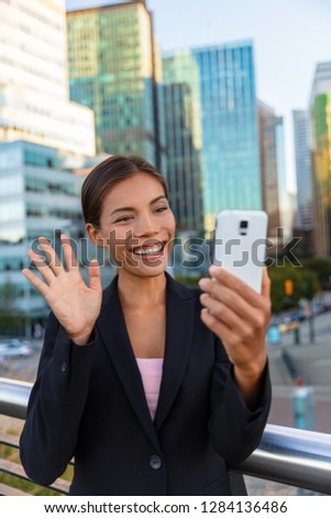 People on video call saying hello waving at camera. Asian woman video calling business partner on smartphone online in city street - Office buidlings on videochat call. Selfie entrepreneur cellphone.