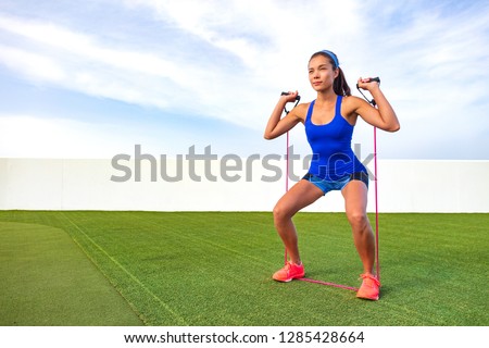Exercise woman with fitness elastic band workout in outdoor gym . Asian girl doing resistance training outside on grass. Fit people lifestyle. Squat and shoulder press with elastics.