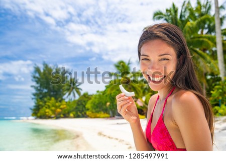 Fresh coconut slice Asian woman eating healthy snack on beach holiday. Summer vacation in Tahiti smiling girl holding fruit cut by local at tropical travel destination. Coco water, popular food trend.