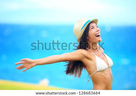 Freedom - free young woman happy on beach smiling joyful and cheerful with arms out on blue ocean sea background. Multicultural woman wearing beach hat and bikini.