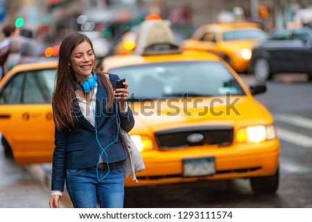 Young woman walking in New York city using phone app for taxi ride hailing with headphones commuting from work. Asian girl happy texting on smartphone. Urban walk commuter NYC.
