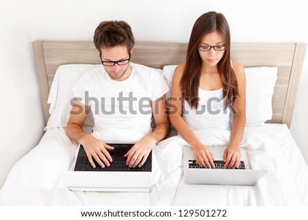 Serious young couple wearing glasses sitting side by side working on their laptops in bed. Beautiful young interracial couple, Asian woman, Caucasian man.