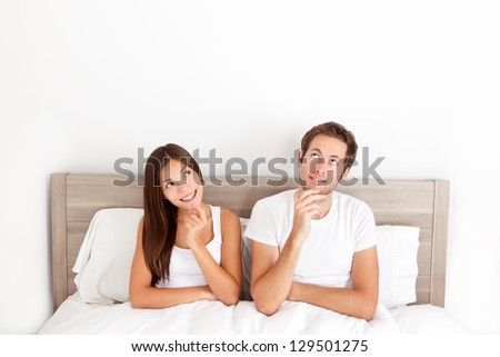 Thinking Young Couple Sitting Pensive In Bed Smiling And Looking Up At The Ceiling For Inspiration And Ideas. Happy Young Interracial Couple, Asian Woman, Caucasian Man.