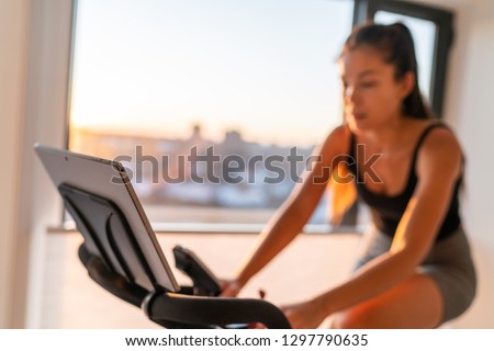 Home fitness fit woman exercising on smart stationary bike at home gym class watching screen online class biking exercise. Young girl training spinning the pedals pedaling. Focus on screen.