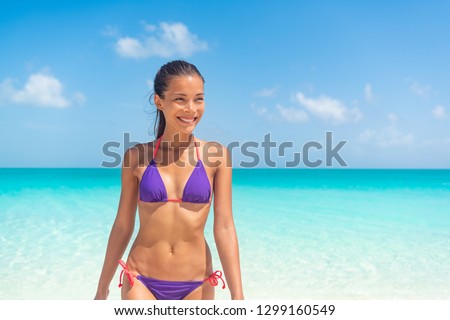 Beautiful Asian fit bikini body model relaxing on Caribbean beach enjoying tropical holidays. Smiling young woman walking on beach.