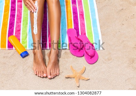 Sunscreen woman applying sun tan cream sunblock lotion on legs with bottle, flip flops and sea star on colorful beach towel on sand. Suntan protection on beach summer travel vacation. female model.