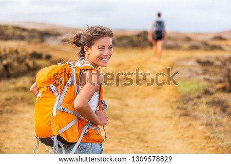 Happy young Asian hiker girl hiking with friend in mountain nature trail wearing orange backpack smiling looking back enjoying travel holiday in summer. Adventure wanderlust lifestyle. Healthy woman.