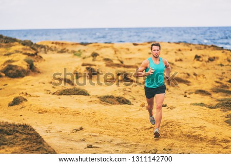 Running man male runner athlete cross country running on golden sand coast by ocean. Young men jogging training on trail outdoors.