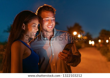 Happy couple using mobile phone at night on city street, faces lit by phone light searching online for restaurant to go out. Smartphone people lifestyle dark background.