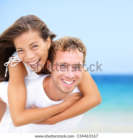 Happy couple on beach fun summer vacation. Multiracial Young newlywed couple piggybacking smiling joyful elated in happiness concept on tropical beach with blue water, sky. Asian woman, Caucasian man.