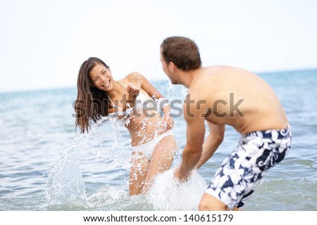 Beach summer fun couple playful splashing water together laughing playing during summer holidays vacation on tropical beach. Beautiful young interracial multiracial couple, Asian woman, Caucasian man.