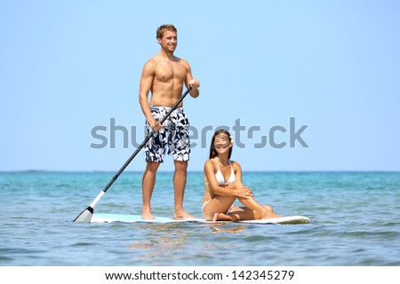 Beach Fun Couple On Stand Up Paddleboard Surfboard Surfing Together In Ocean Sea On Big Island, Hawaii Beautiful Young Multi-Ethnic Couple, Mixed Race Asian Woman And Caucasian Man Doing Water Sport.