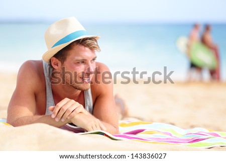 Man On Beach Lying In Sand Looking To Side Smiling Happy Wearing Hipster Summer Hat. Young Male Model Enjoying Summer Travel Holiday By The Ocean.