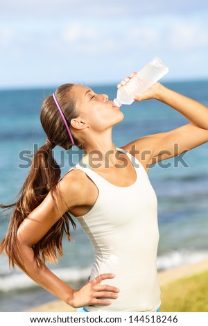 Fitness woman drinking water after running at beach. Thirsty sport runner resting taking a break with water bottle drink outside after training. Beautiful fit sporty mixed race Asian girl.